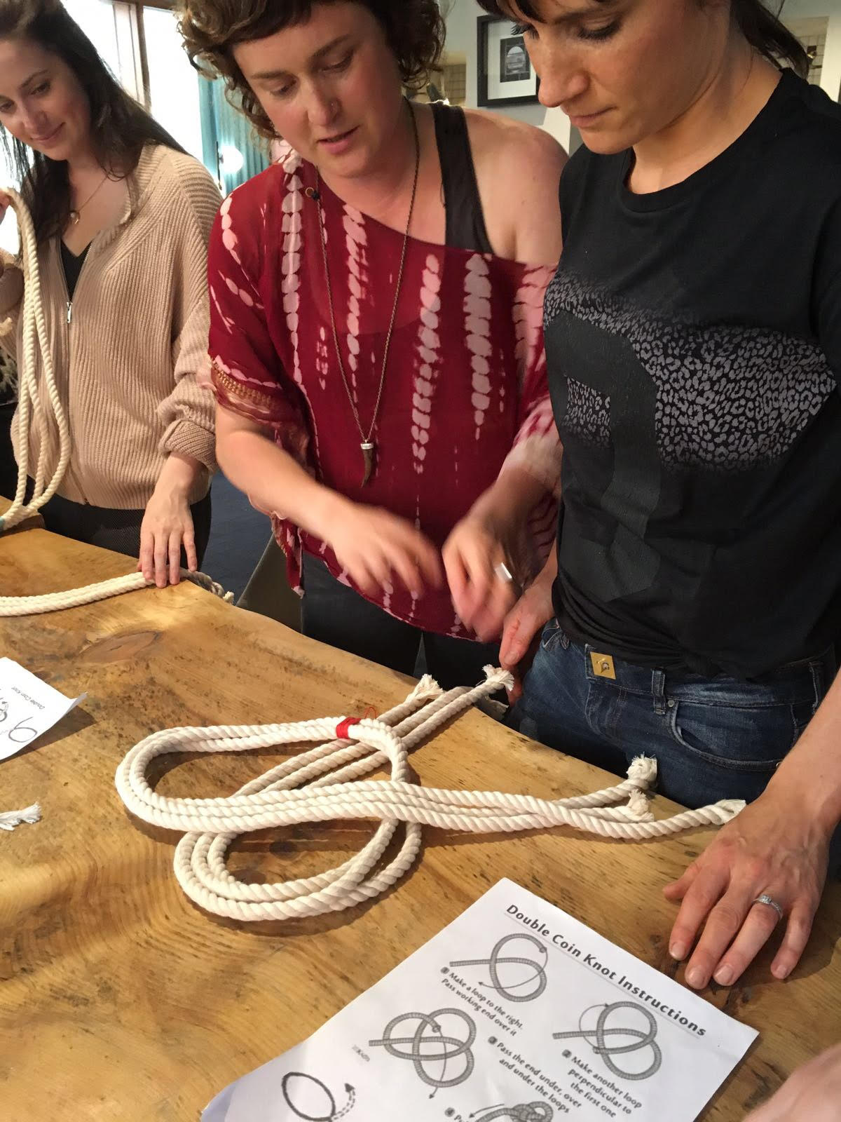 Two women working with a group of ropes on a wooden table, with a knot instruction sheet in the foreground and another person watching in the background