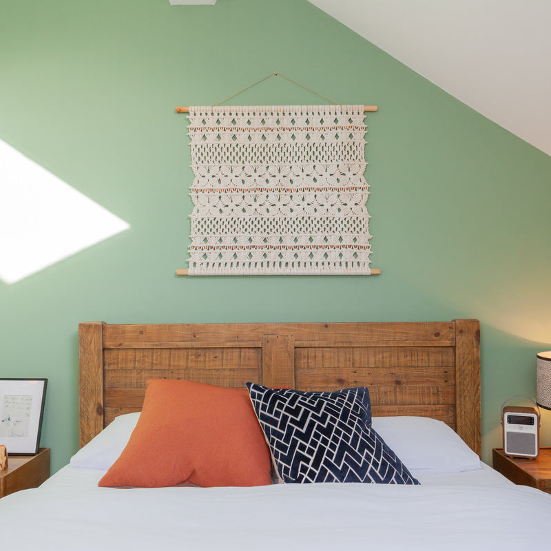 An ASTRL Fibres macrame tapestry styled against a light green wall, above a bed with 2 patterned pillows. 