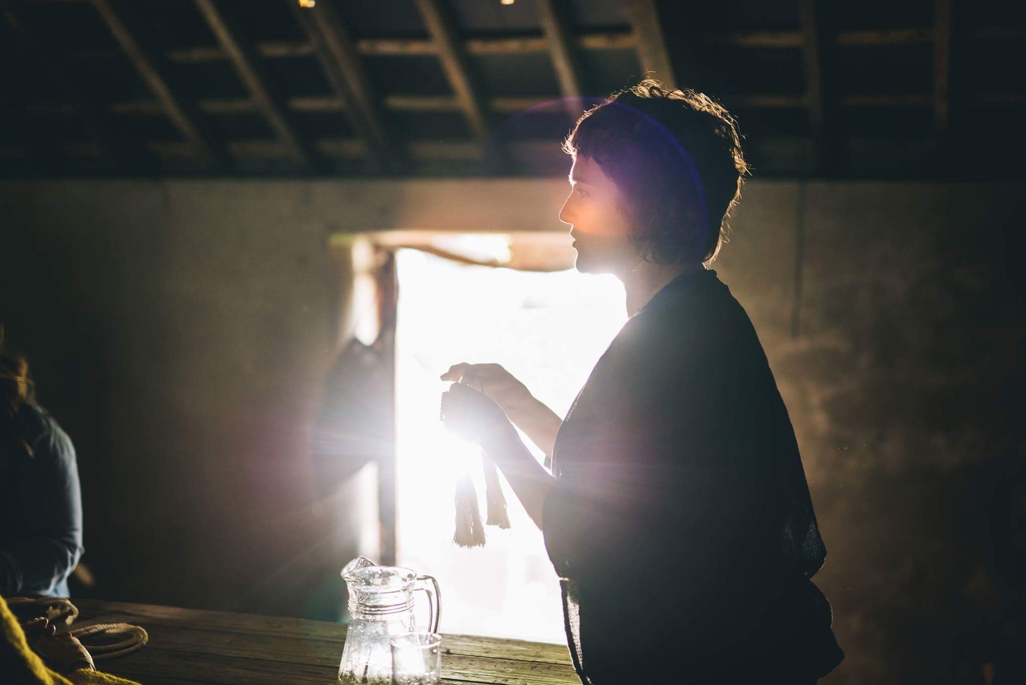 A profile of a woman in front of a bright window in a rustic room with wooden rafters holding fibres
