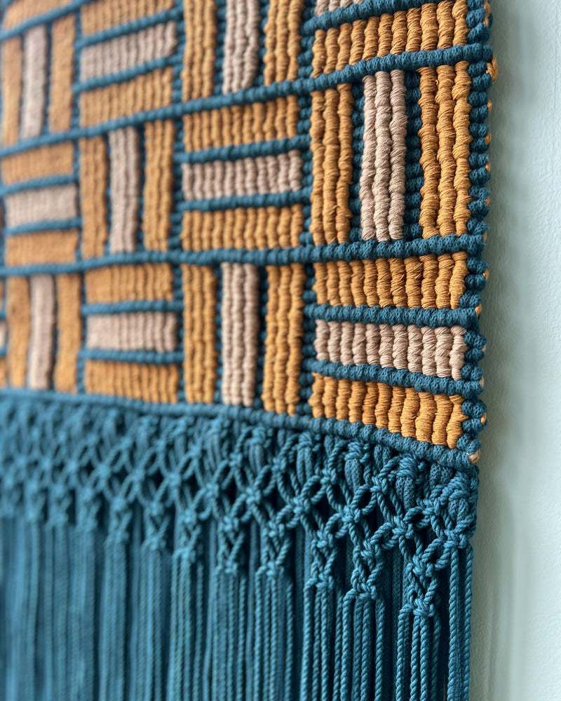 A close up of a macrame tapestry with a geometric tile pattern in blue and 2 shades of brown against a light blue wall