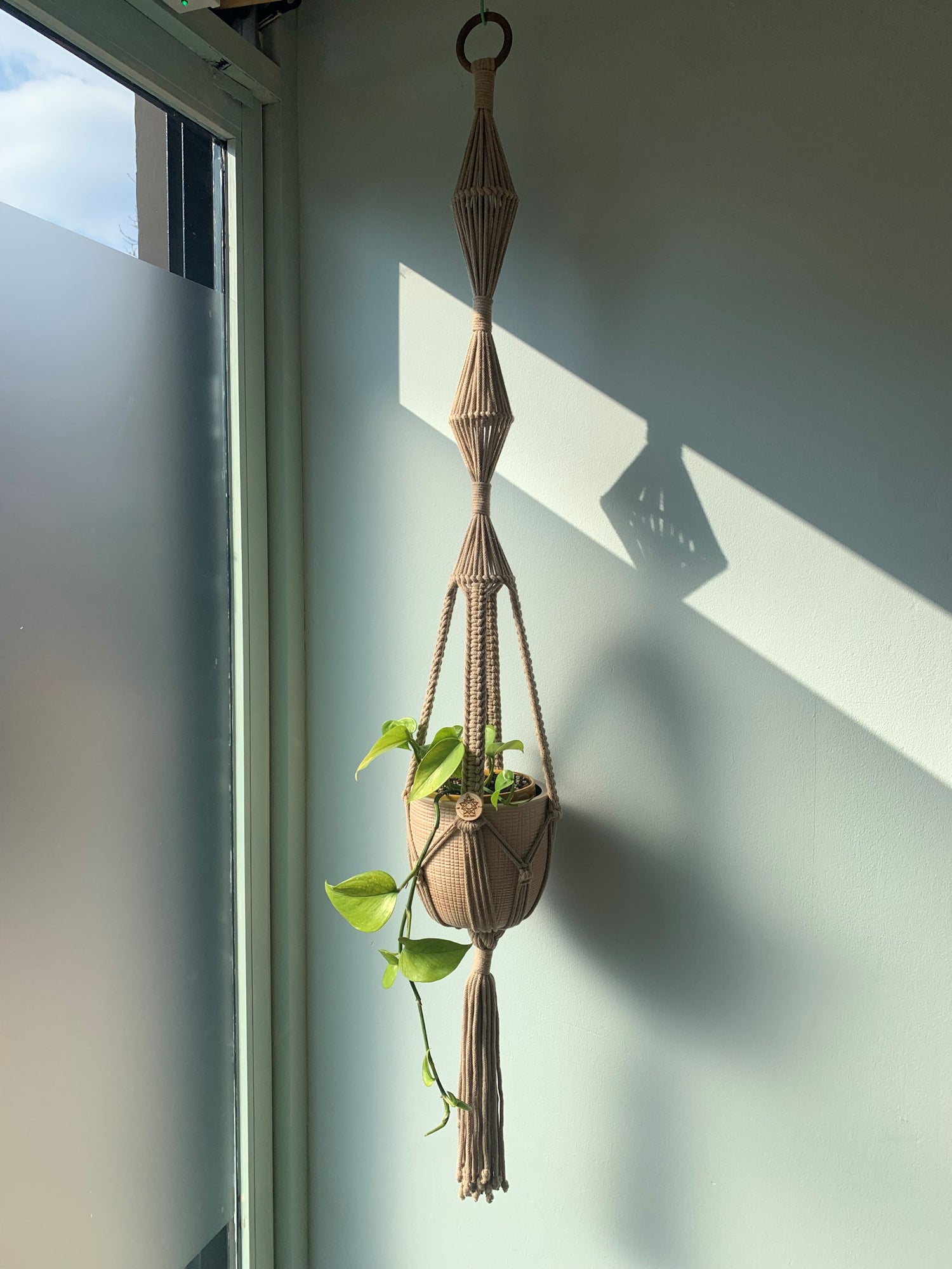 A modern plant hanger in taupe with a zig zag design hanging by a sunny window casting shadows on a light blue wall