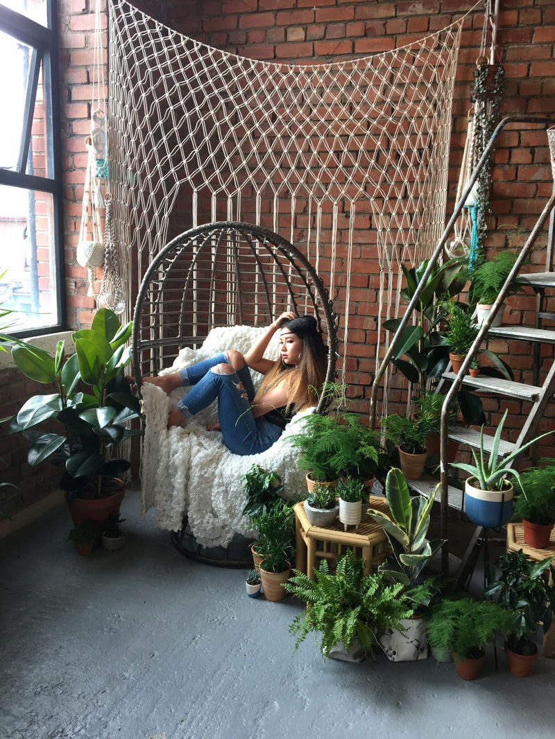 A woman lounging in a wicker chair by a window, surrounded by plants and a macrame backdrop