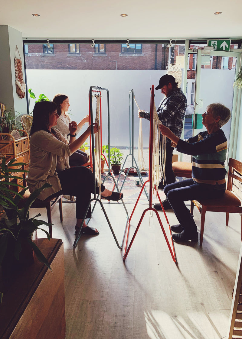 4 people sitting and standing in front of rails during a macramé plant hanger workshop in the light filled ASTRL Fibres Studio in Belfast. 