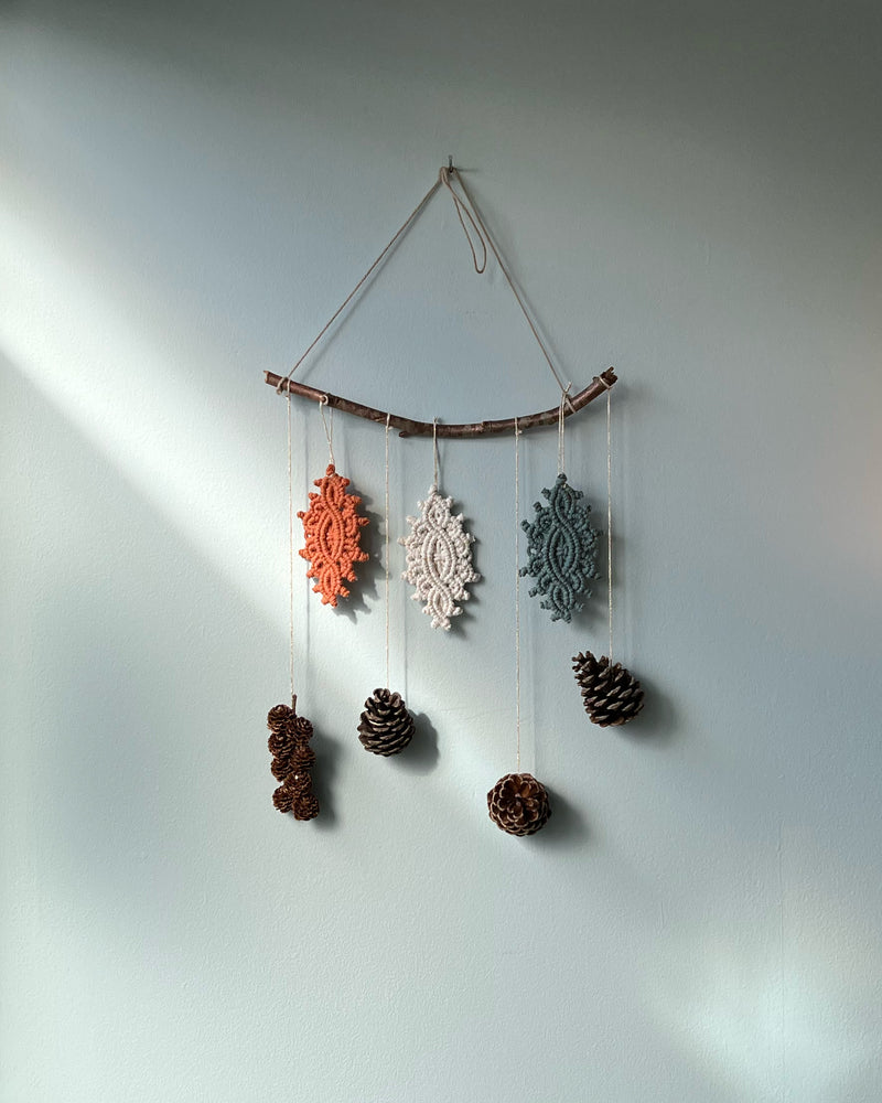 A hanging branch with 3 intricate macrame ornament and pine cones hung on it against a light blue wall