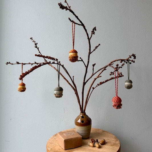 Traditional passementerie acorn baubles hanging on branches placed in a vase against a light blue wall.