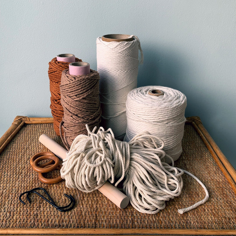 Spools of yarn, wooden rings, a dowel and S hooks on a woven rattan table against a light blue wall.