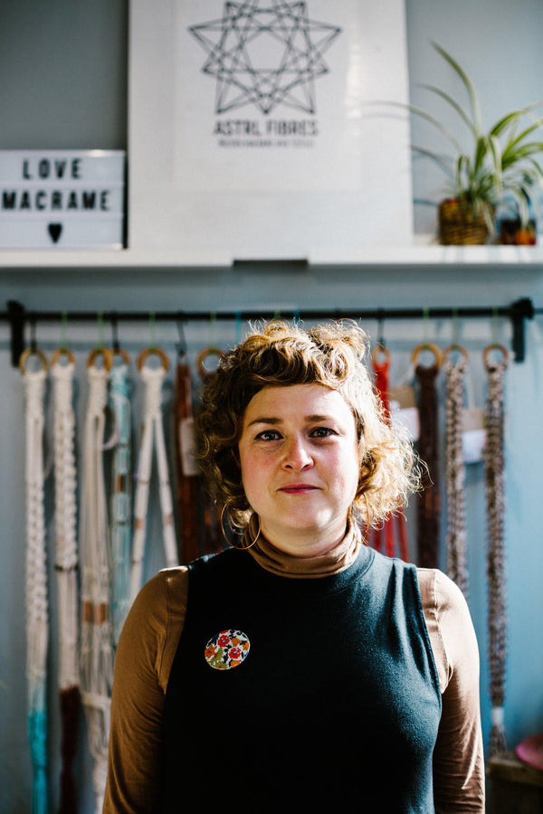 Woman standing in front of a rail on a light blue wall with macrame items and 'ASTRL Fibers' branding in the background.