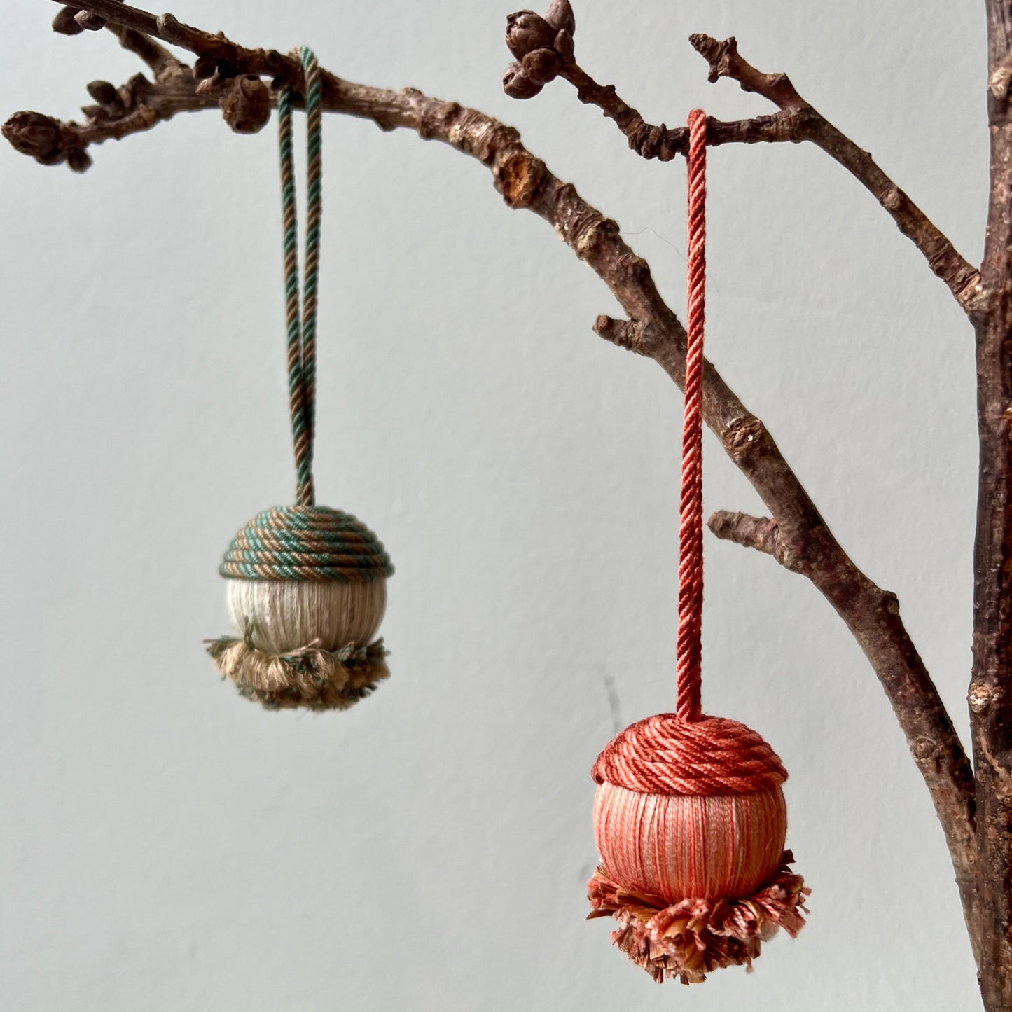 Two passementerie acorn ornaments hanging from a branch against a light blue background