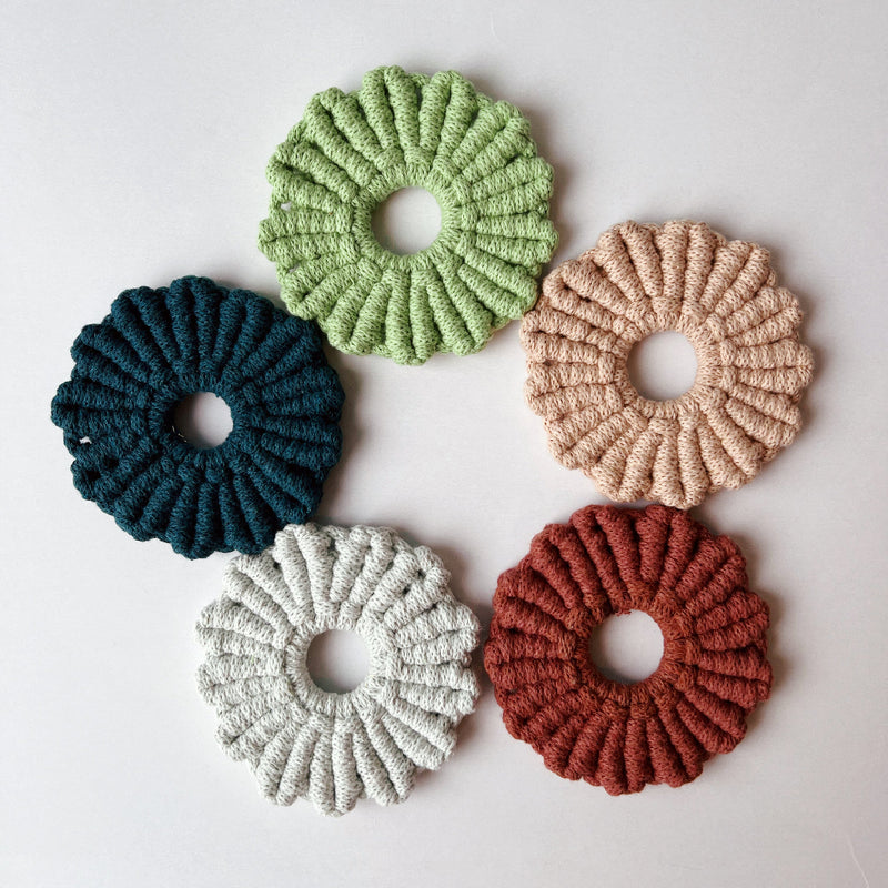 A set of five knotted daisy coasters in blue, white, green, blush, and rust in a circle on a white background