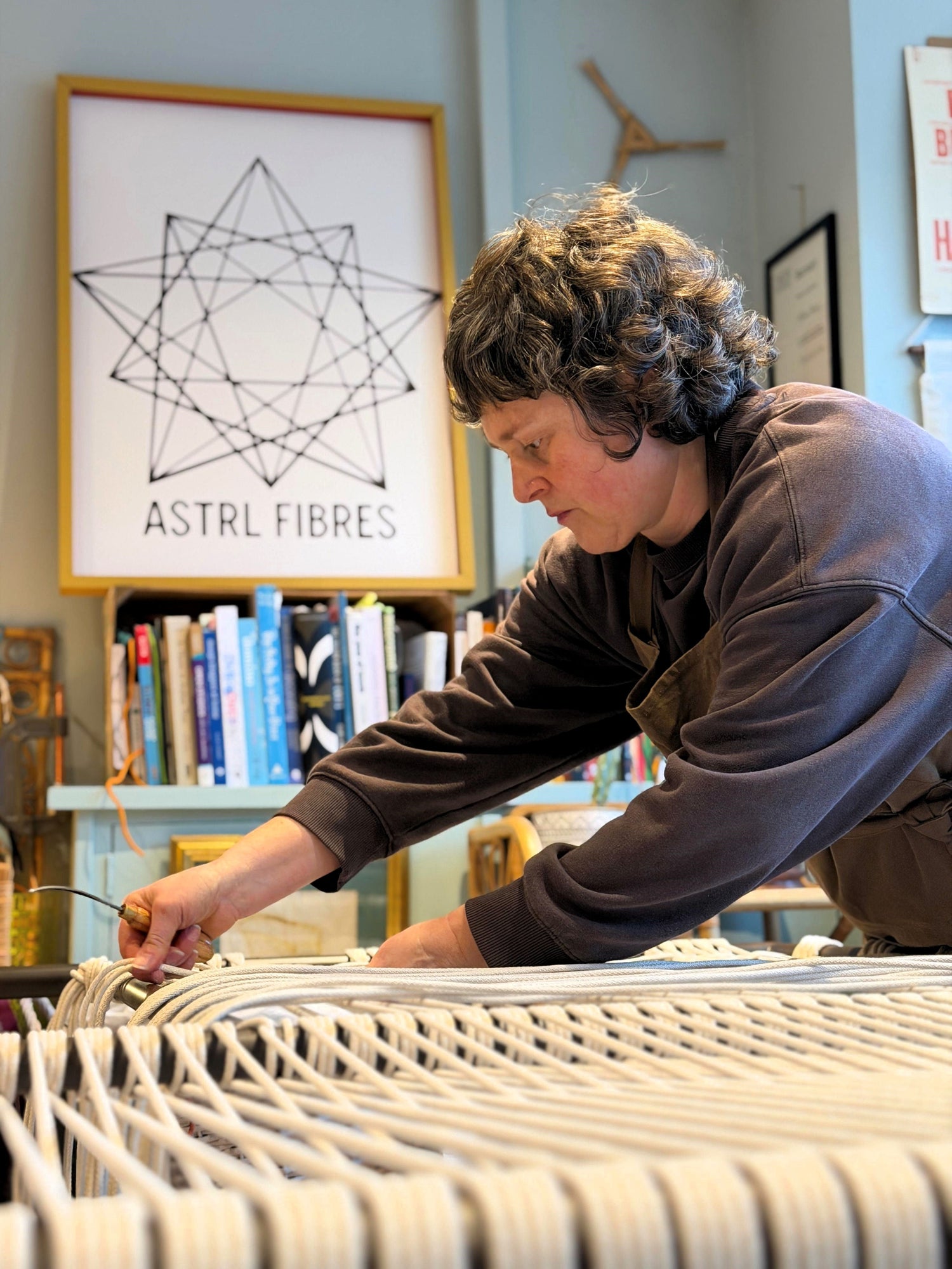 Woman working on a woven screen in a room with books and a framed picture with the ASTRL Fibres logo in the bakground