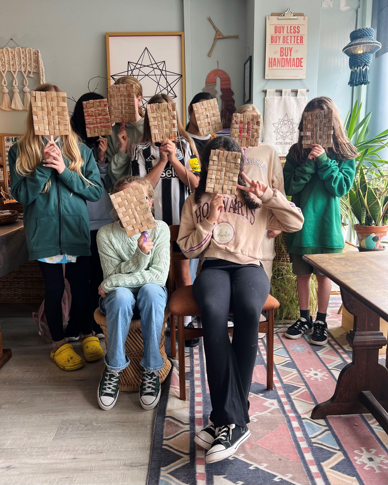 9 young people surrounded by plants and macramé artwork, holding handmade paper fans in front of their faces sitting in the ASTRL Fibres studio in Belfast.