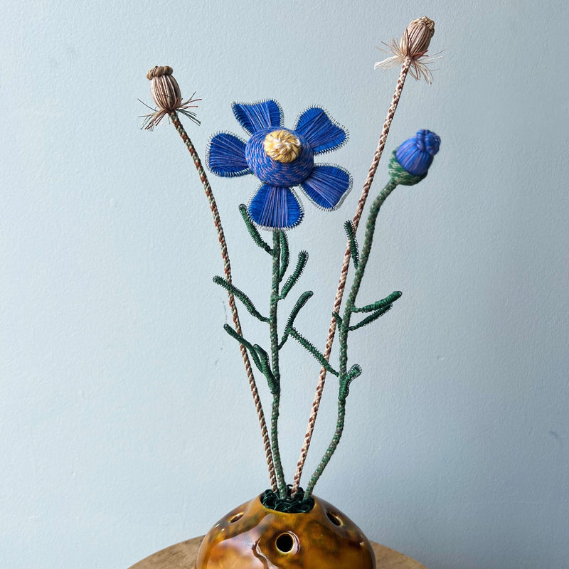 Passementerie design of a blue & yellow flower with green leaves and Kumihimo braided stem and natural buds and stems on a light blue background places in a brown vase