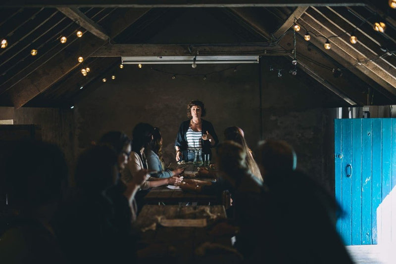 Alison Pascoe standing at the top of a table teaching a traditional knot workshop in a converted barn strung with fairy lights