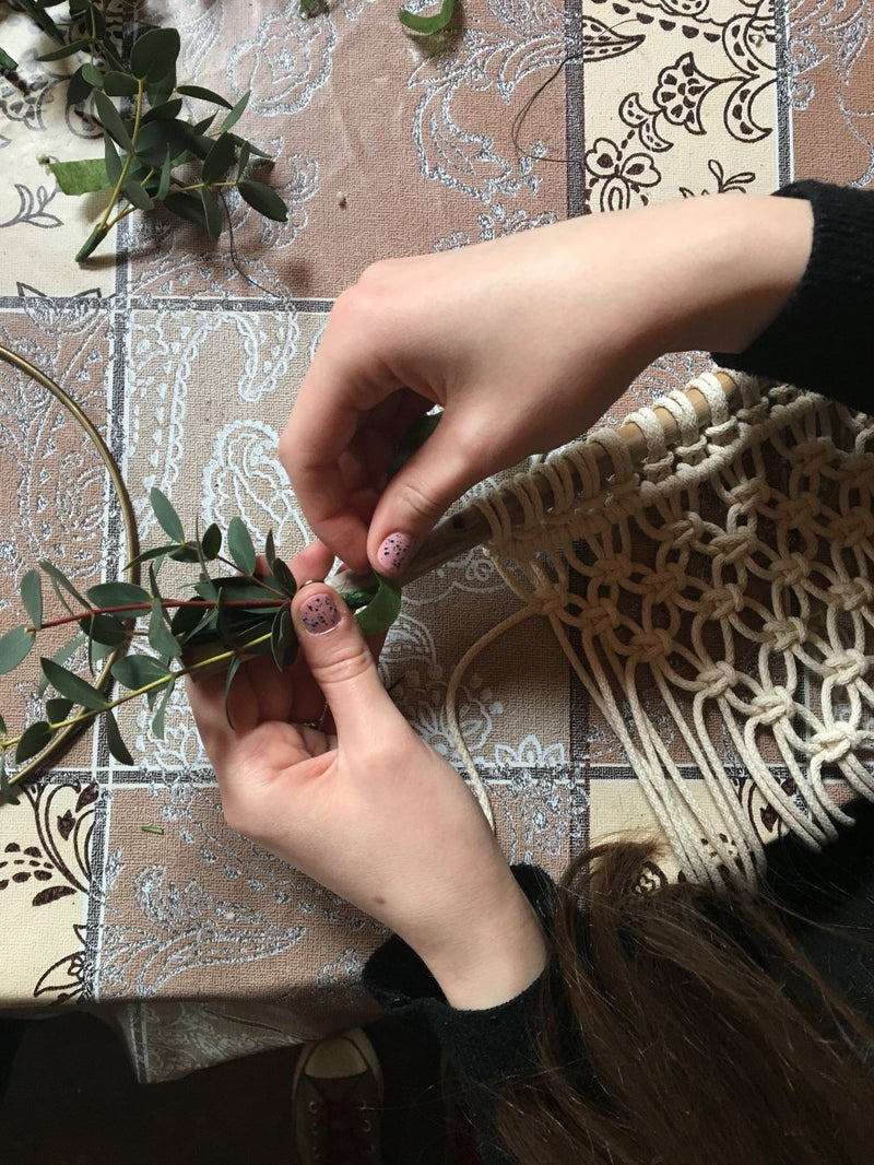 A detail of a woman's hands adding eucalyptus leaves to a handmade macramé wall hanging above a tiled table top.