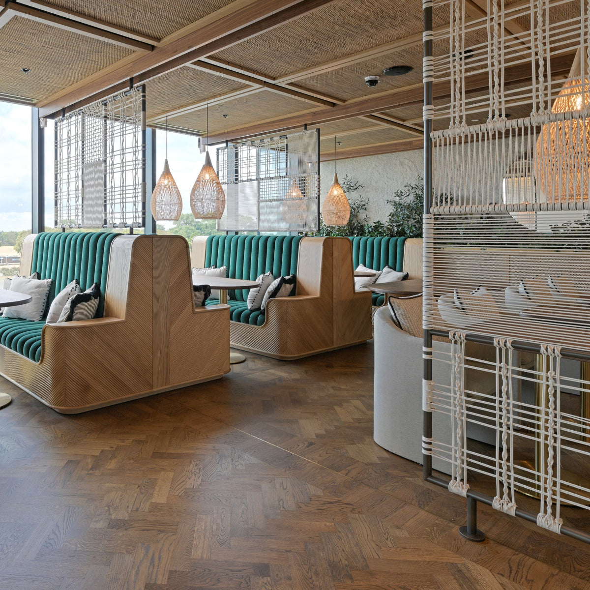 A modern indoor seating area with handcrafted wooden booth chairs, teal upholstered benches, pendant lights, woven screen dividers and a large window looking out onto the river Thames and the landscape beyond.
