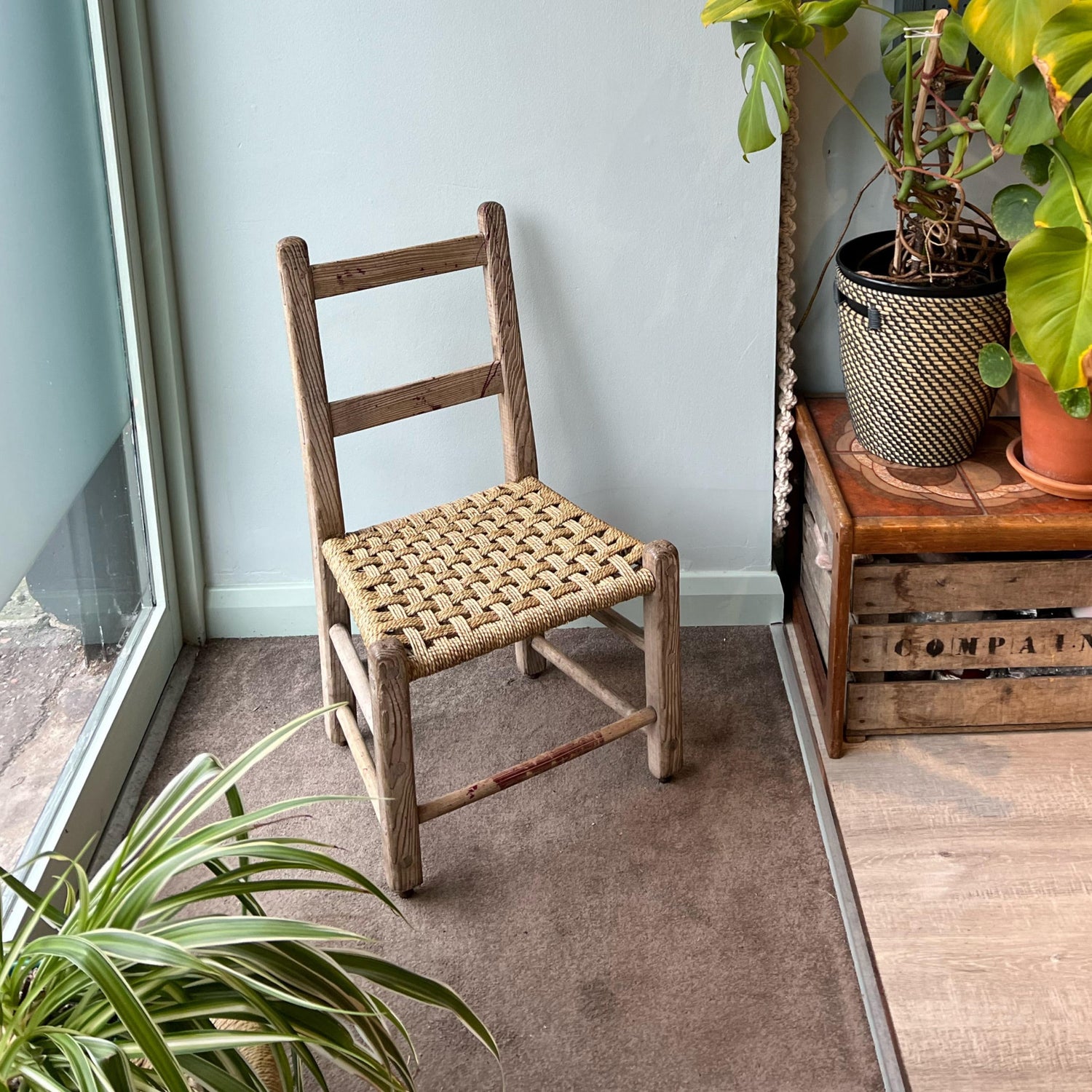 Rustic wooden chair with woven seat in a room with plants and a window.